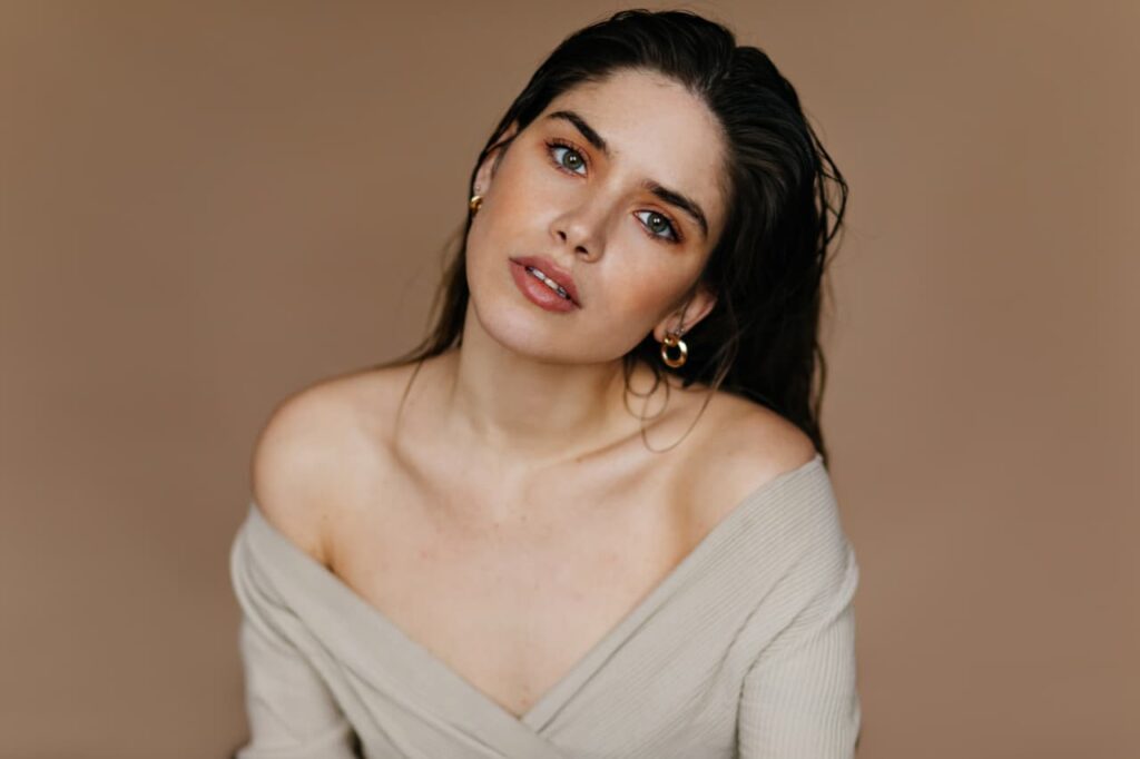 a women with tilted head aestheticely posing wearing some subtle makeup with some minimal earrings and beige coloured top